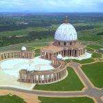 Vue aérienne de la " basilique de Yamoussoukro", un grand bâtiment en forme de dôme avec des structures environnantes plus petites, au milieu de vastes pelouses vertes et d'arbres sous un ciel partiellement nuageux en Côte d'Ivoire.