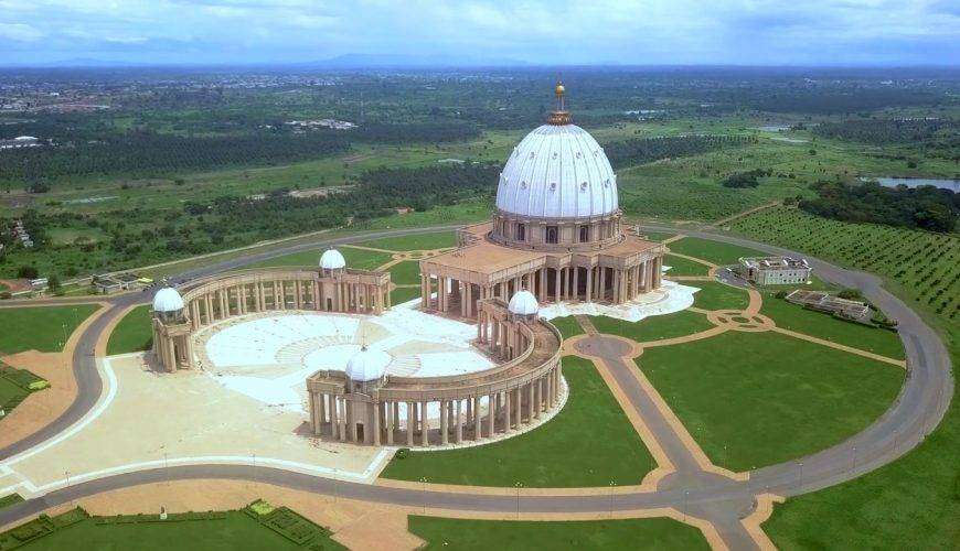 Vue aérienne de la " basilique de Yamoussoukro", un grand bâtiment en forme de dôme avec des structures environnantes plus petites, au milieu de vastes pelouses vertes et d'arbres sous un ciel partiellement nuageux en Côte d'Ivoire.