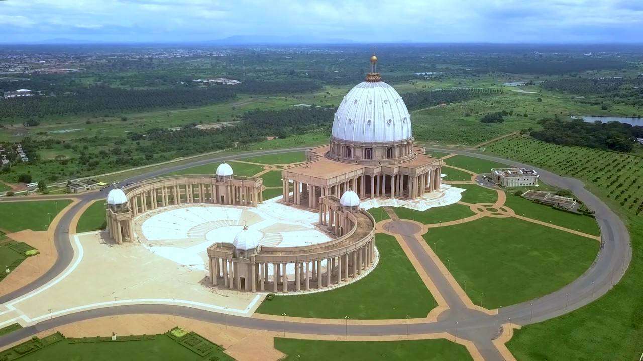 Vue aérienne de la " basilique de Yamoussoukro", un grand bâtiment en forme de dôme avec des structures environnantes plus petites, au milieu de vastes pelouses vertes et d'arbres sous un ciel partiellement nuageux en Côte d'Ivoire.