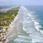 Une vue aérienne sereine de la côte tropicale de la Côte d'Ivoire avec des vagues qui clapotent doucement contre le rivage, bordées de palmiers luxuriants et parsemées de bâtiments, soulignant la rencontre tranquille de la terre.