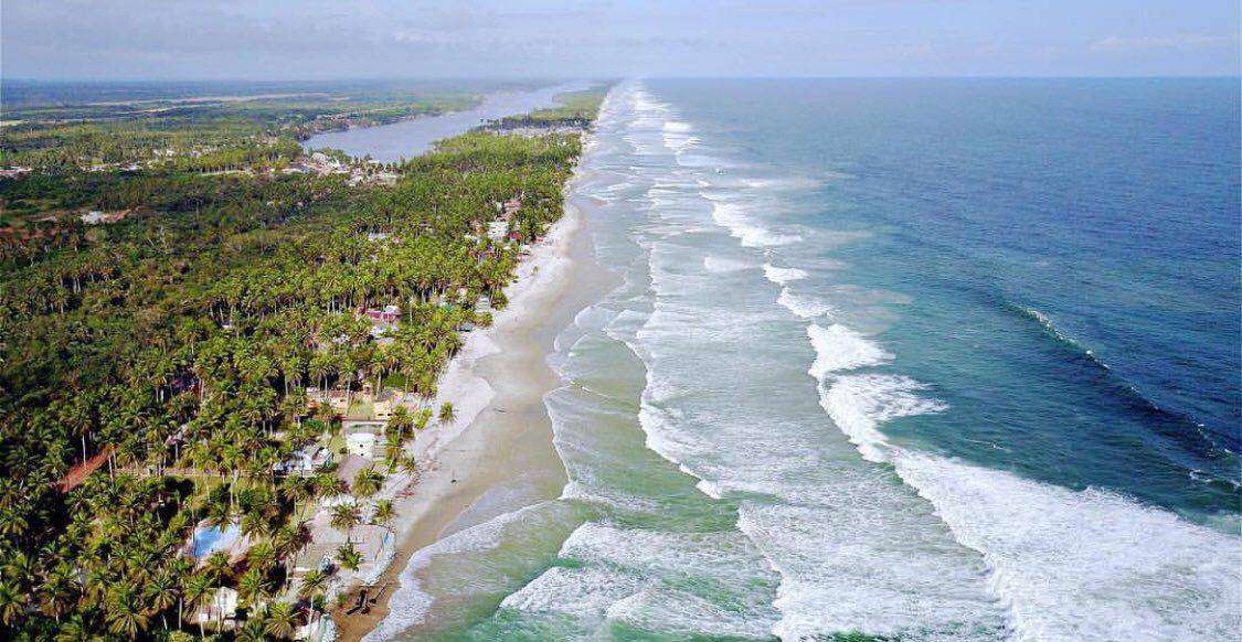 Une vue aérienne sereine de la côte tropicale de la Côte d'Ivoire avec des vagues qui clapotent doucement contre le rivage, bordées de palmiers luxuriants et parsemées de bâtiments, soulignant la rencontre tranquille de la terre.