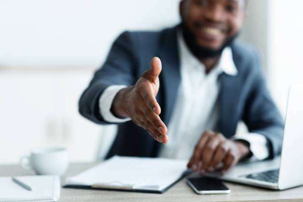 Un homme d'affaires souriant tendant la main pour une poignée de main à son bureau, avec un ordinateur portable, un smartphone et des documents en vue, signalant un accueil amical ou la conclusion d'une transaction en Côte d'I