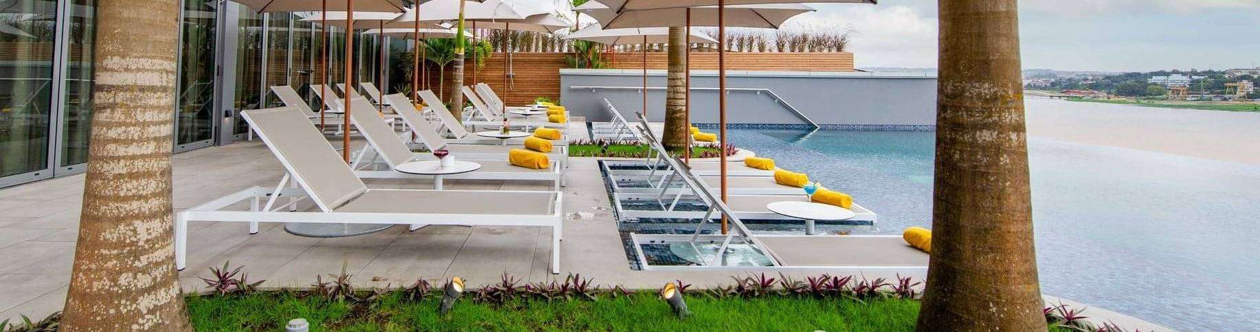 Une retraite tranquille au bord de la piscine en Côte d'Ivoire avec des chaises longues et des parasols modernes sur fond d'eaux calmes et de verdure luxuriante, offrant une évasion sereine pour la détente et les loisirs.