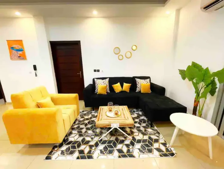 Salon moderne avec un superbe canapé noir en forme de L rehaussé de coussins jaunes, un fauteuil jaune moutarde et un tapis à motifs ancrant une élégante table basse en bois. L'espace est plus loin