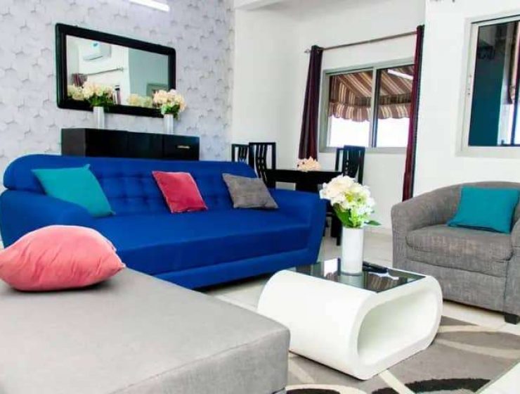 Salon lumineux et contemporain avec un canapé bleu orné de coussins colorés, complété par un fauteuil gris, une table basse blanche et un élégant coin repas inspiré de la Côte d'Ivoire