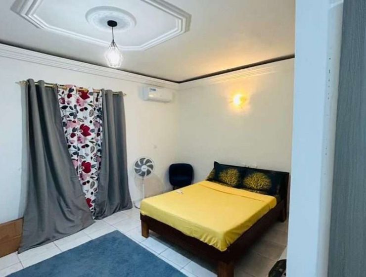 Une chambre confortable dotée d'un lit simple avec une literie jaune d'inspiration ivoirienne, des rideaux à motifs, un ventilateur de plafond et un éclairage tamisé créant une ambiance confortable.