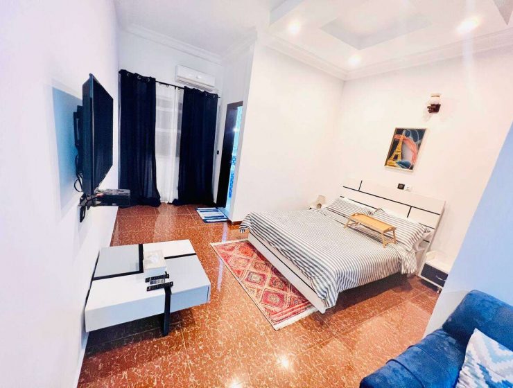 Une chambre moderne et soignée avec des murs blancs, un sol en terre cuite à motifs inspirés de la Côte d'Ivoire, un couvre-lit à rayures noires et blanches et un mobilier contemporain dont un noir.